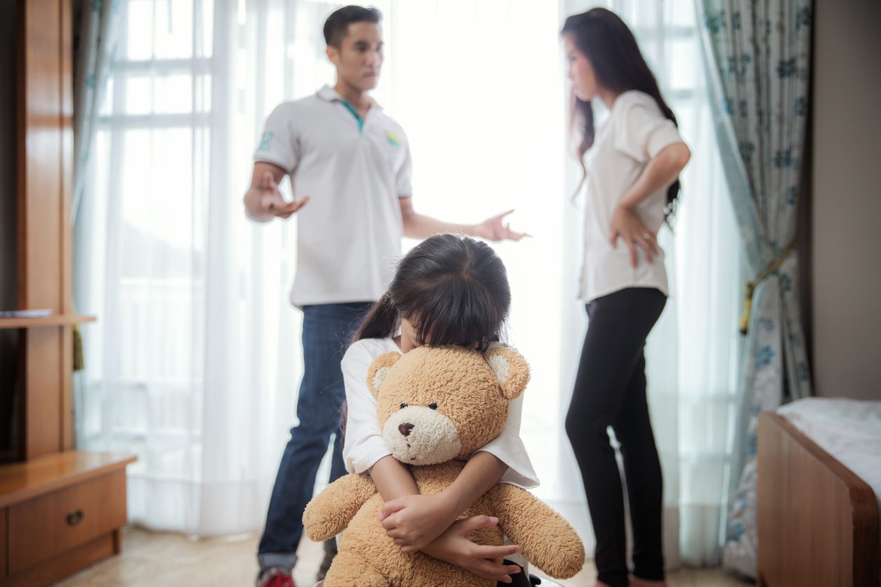 A girl holding onto a teddy bear while two people watch.